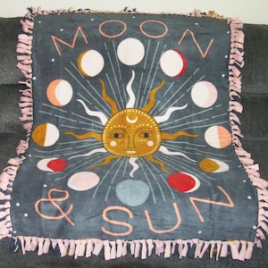 May include: A gray fleece blanket with a pink fringe border. The blanket features a sun and moon design with the text "Moon & Sun".