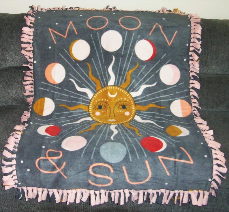 May include: A gray fleece blanket with a pink fringe border. The blanket features a sun and moon design with the text "Moon & Sun".