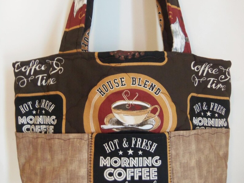 Puede incluir: Un bolso tote acolchado con un estampado de caf&eacute; en tonos marr&oacute;n y negro. La tela muestra textos como "Coffee Time", "House Blend" y "Hot & Fresh Morning Coffee" junto a ilustraciones de tazas de caf&eacute; con vapor sobre un fondo oscuro.