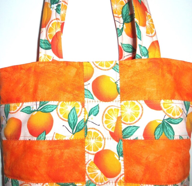 May include: A handmade tote bag with a vibrant orange and white citrus fruit pattern. The bag features a patchwork design with solid orange panels and handles, and a repeating pattern of oranges and green leaves. The bag is designed for carrying items.