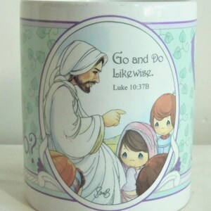 May include: A white ceramic mug featuring a central oval illustration of a bearded figure in a white robe pointing toward two children. The text "Go and Do Likewise. Luke 10:37B" is printed in black. The background has a light green ivy pattern.