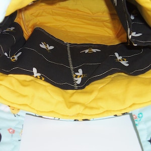 K&ouml;nnte beinhalten: Das Innere einer gesteppten Stofftasche mit leuchtend gelbem Futter und einer schwarzen Tasche mit wiederkehrendem Bienenmuster. Der Au&szlig;enstoff ist hellblau mit Bienen- und Blumenmuster und enth&auml;lt ein wei&szlig;es, rechteckiges Etikett.