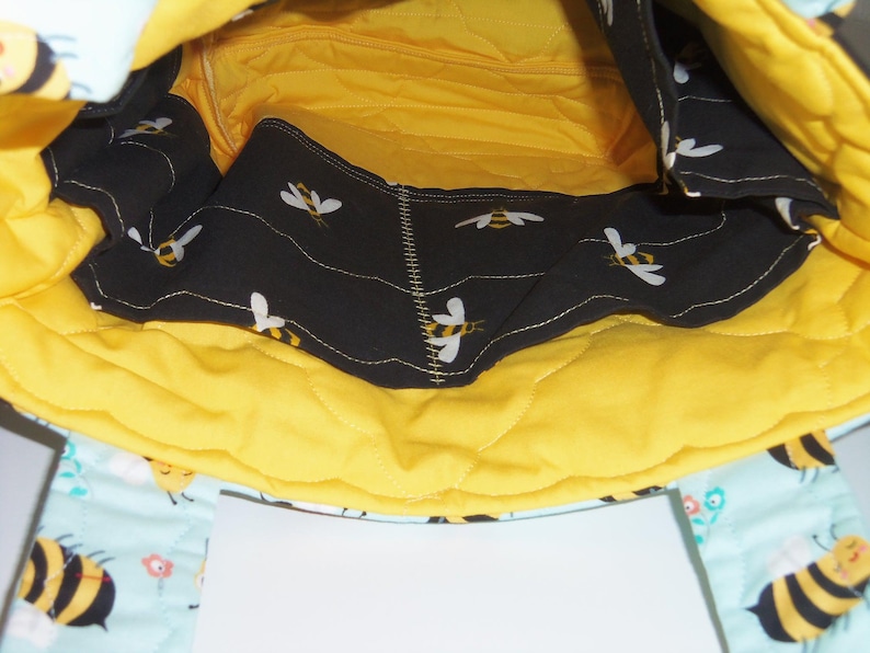 K&ouml;nnte beinhalten: Das Innere einer gesteppten Stofftasche mit leuchtend gelbem Futter und einer schwarzen Tasche mit wiederkehrendem Bienenmuster. Der Au&szlig;enstoff ist hellblau mit Bienen- und Blumenmuster und enth&auml;lt ein wei&szlig;es, rechteckiges Etikett.