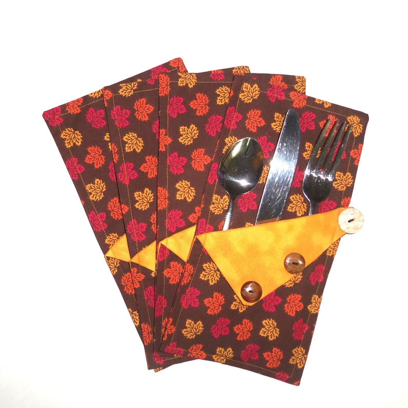 May include: Set of four brown fabric placemats with a pattern of orange and red maple leaves. The placemats have a yellow pocket to hold a fork, knife, and spoon.