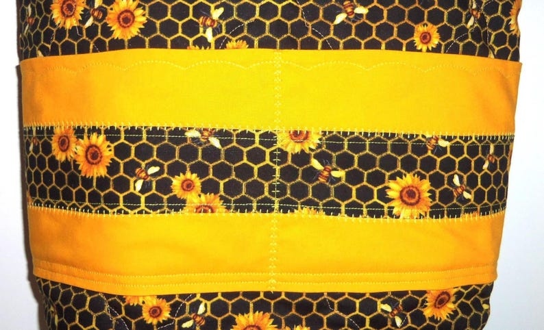 May include: A quilted fabric item featuring a black background with a yellow honeycomb pattern, sunflowers, and bees. The design includes horizontal solid yellow bands with scalloped edges and decorative stitching across the surface.
