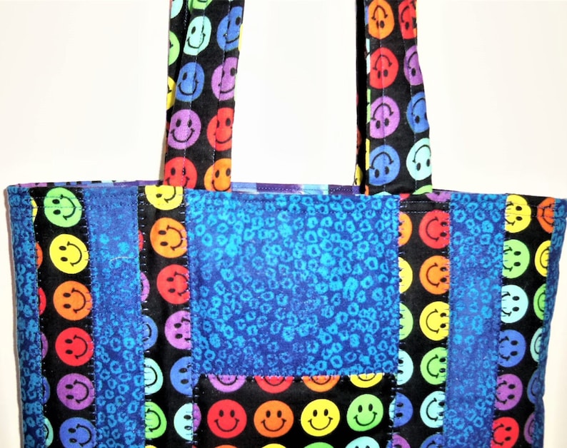 Puede incluir: Un bolso de tela con un dise&ntilde;o de patchwork de tela negra estampada con caras sonrientes de colores y tela azul con un patr&oacute;n animal texturizado. El bolso incluye dos asas de tela a juego con el mismo estampado de caras sonrientes.