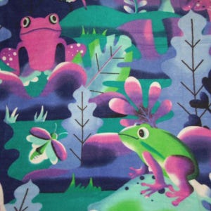 May include: A fleece blanket with a playful frog and butterfly pattern. The blanket has a colour scheme of green, pink, purple, and blue, with a fringed edge. The design includes stylised plants and mushrooms.