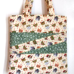 K&ouml;nnte beinhalten: Eine gesteppte Tragetasche aus cremefarbenem Stoff mit einem sich wiederholenden Muster aus Katzen, die verschiedene Accessoires wie Zylinder, Baskenm&uuml;tzen und Blumenkr&auml;nze tragen, akzentuiert durch zwei diagonale B&auml;nder aus gr&uuml;nem Stoff.