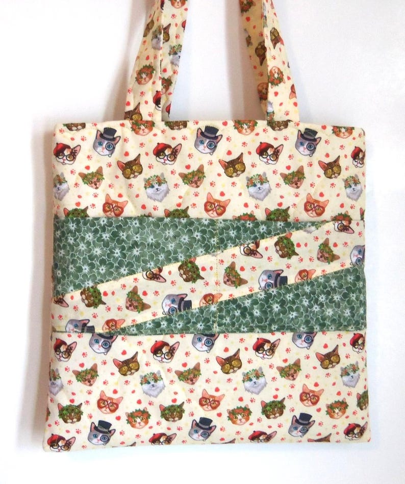 K&ouml;nnte beinhalten: Eine gesteppte Tragetasche aus cremefarbenem Stoff mit einem sich wiederholenden Muster aus Katzen, die verschiedene Accessoires wie Zylinder, Baskenm&uuml;tzen und Blumenkr&auml;nze tragen, akzentuiert durch zwei diagonale B&auml;nder aus gr&uuml;nem Stoff.