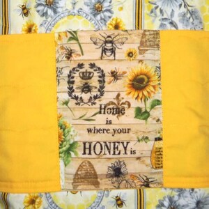 May include: A quilted fabric item with a yellow band and a central panel featuring bees, sunflowers, and the text "Home is where your HONEY is." The background has a floral and honeycomb pattern.