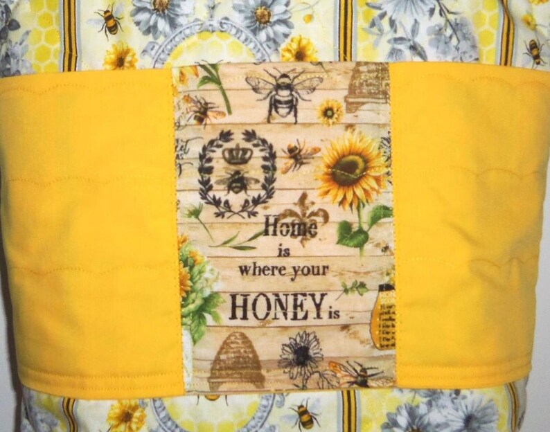May include: A quilted fabric item with a yellow band and a central panel featuring bees, sunflowers, and the text "Home is where your HONEY is." The background has a floral and honeycomb pattern.