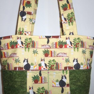 May include: A quilted tote bag featuring a light yellow fabric printed with a pattern of cats, potted plants, and books on shelves. The bottom section of the bag is made of a solid dark green fabric with a textured, mottled appearance.