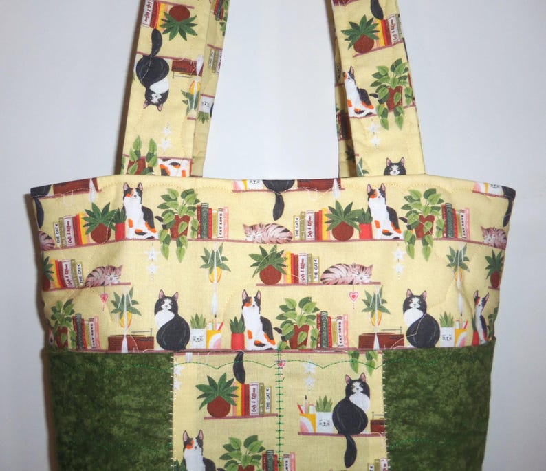 May include: A quilted tote bag featuring a light yellow fabric printed with a pattern of cats, potted plants, and books on shelves. The bottom section of the bag is made of a solid dark green fabric with a textured, mottled appearance.