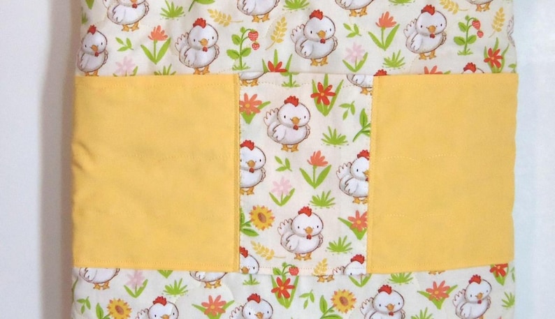 May include: A quilted fabric item featuring a repeating pattern of white chickens with red combs, green grass, and orange and yellow flowers. A solid yellow horizontal band spans the center, dividing the patterned fabric into three sections.