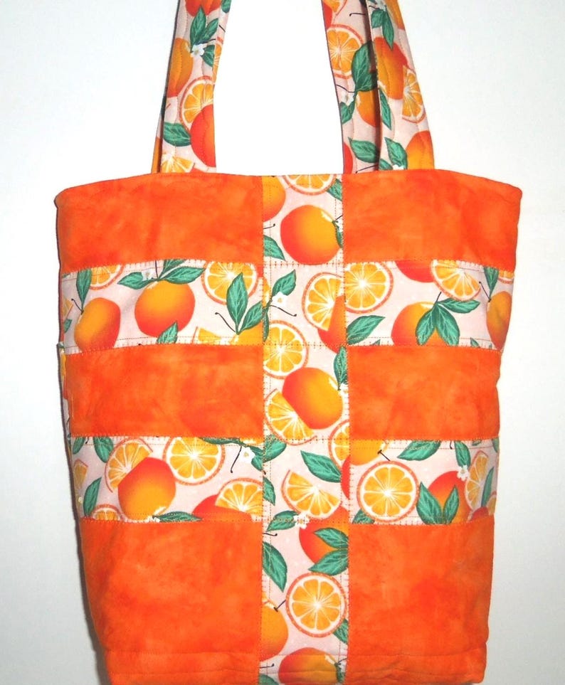 May include: A quilted tote bag with a vibrant orange and white citrus fruit pattern. The bag features solid orange panels and handles, with a repeating design of oranges and orange slices on a white background. The bag is designed for carrying items.