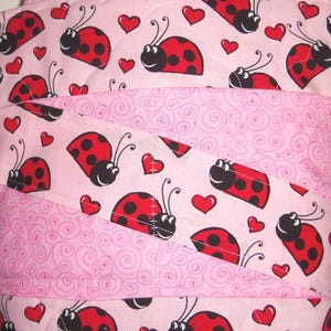 May include: A quilted fabric item featuring a light pink background with a repeating pattern of red and black ladybugs and small red hearts, interspersed with diagonal bands of light pink fabric decorated with a subtle tonal swirl pattern.