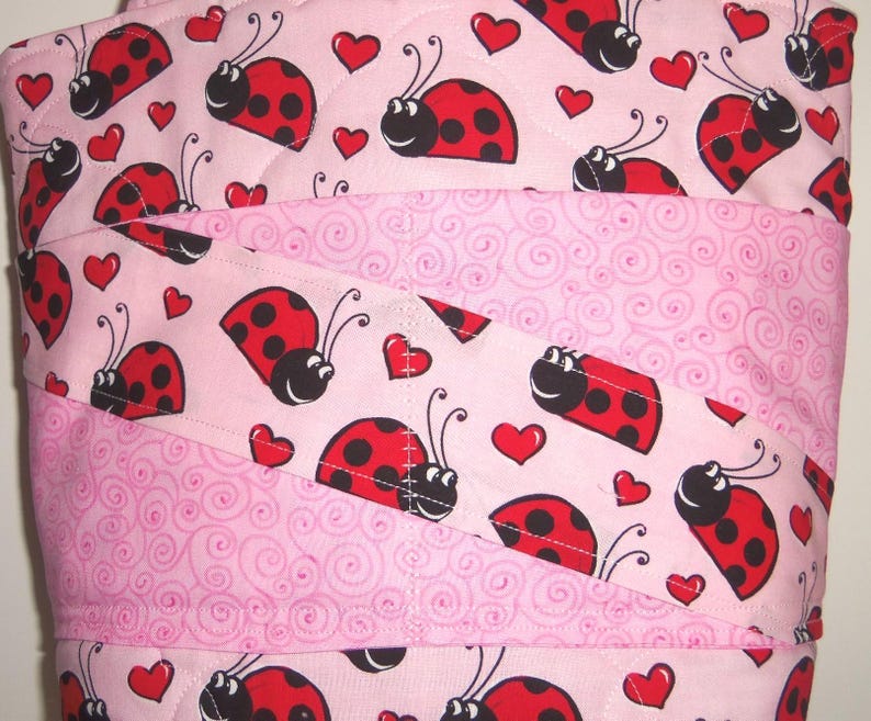 May include: A quilted fabric item featuring a light pink background with a repeating pattern of red and black ladybugs and small red hearts, interspersed with diagonal bands of light pink fabric decorated with a subtle tonal swirl pattern.