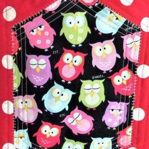 May include: A quilted fabric item featuring a red background with white polka dots and a central pentagonal pocket. The pocket is black with a colourful owl pattern in shades of pink, green, blue, and yellow, including the text snooze and zzz.