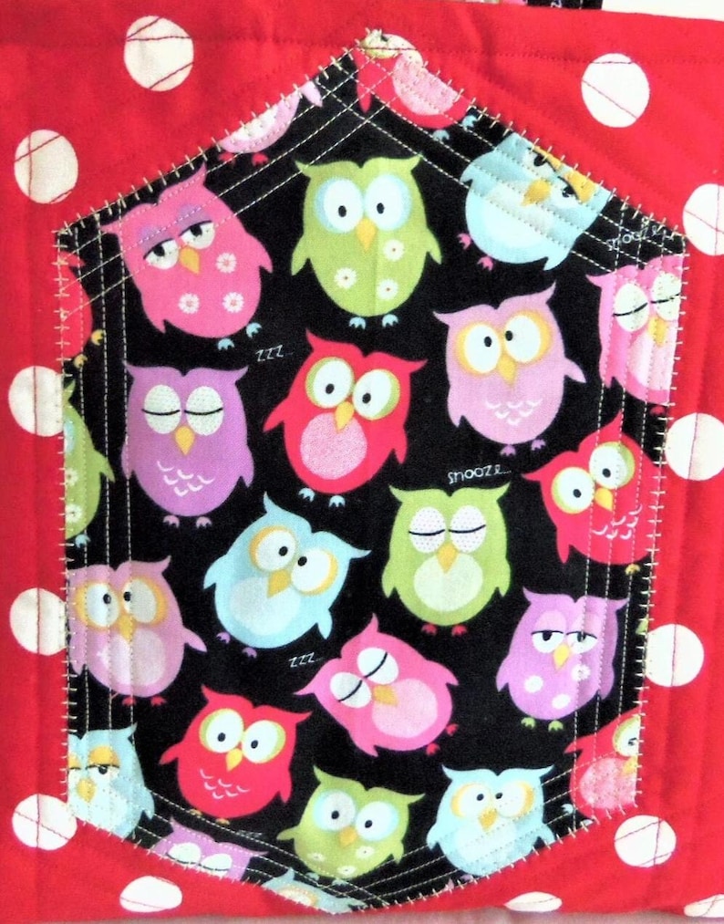 May include: A quilted fabric item featuring a red background with white polka dots and a central pentagonal pocket. The pocket is black with a colourful owl pattern in shades of pink, green, blue, and yellow, including the text snooze and zzz.
