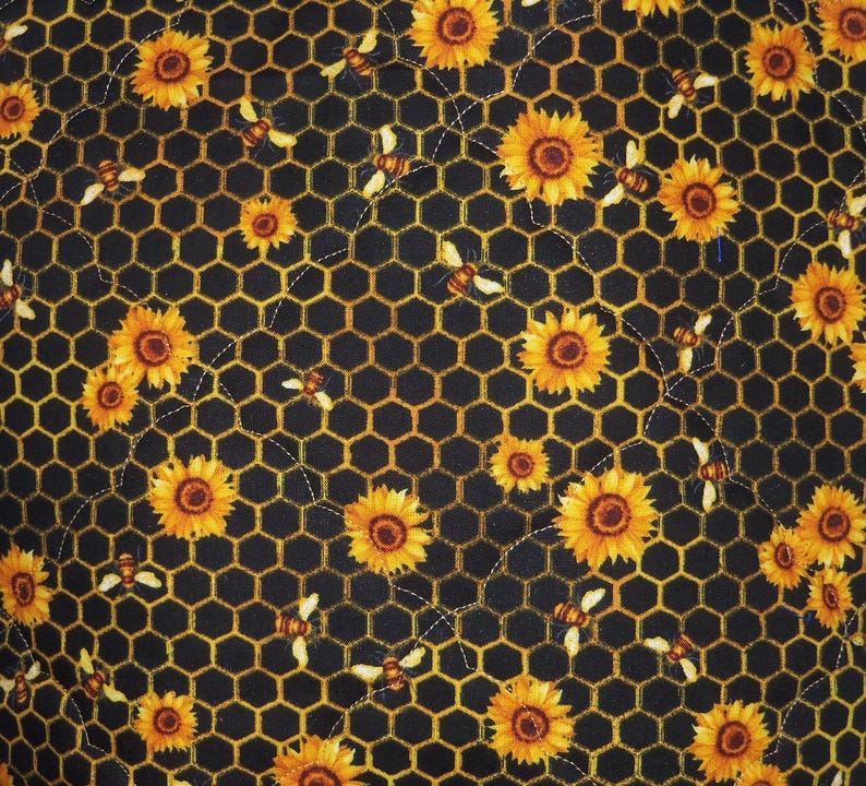 May include: A fabric featuring a repeating pattern of yellow sunflowers and small bees scattered across a black background with a yellow honeycomb grid. The material has visible white stitching lines that create a quilted texture throughout.
