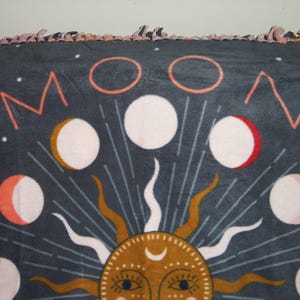 May include: A dark blue fleece blanket with a moon and sun design. The moon phases are depicted in pink, white, and gold. The sun is gold with a crescent moon on its forehead and has a face with eyes and a nose.