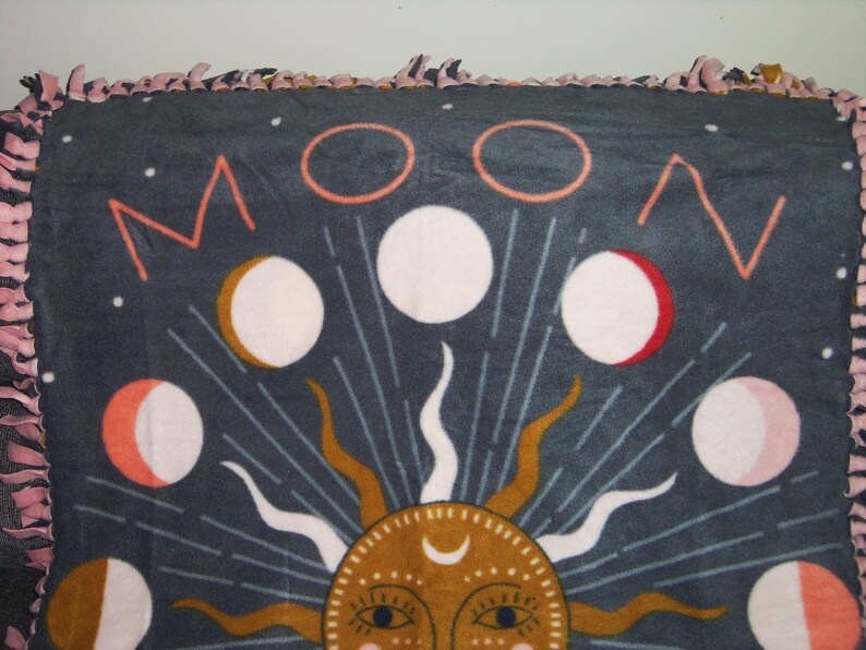 May include: A dark blue fleece blanket with a moon and sun design. The moon phases are depicted in pink, white, and gold. The sun is gold with a crescent moon on its forehead and has a face with eyes and a nose.