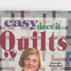 以下が含まれることがあります： ジョージア・ボーンスティール著のハードカバー本「Easy Does It Quilts」。表紙は明るいグリッド模様の背景で、タイトルは大きな紫、緑、赤の文字で書かれており、下部には人物の写真が掲載されています。