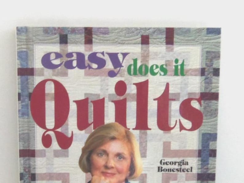 以下が含まれることがあります： ジョージア・ボーンスティール著のハードカバー本「Easy Does It Quilts」。表紙は明るいグリッド模様の背景で、タイトルは大きな紫、緑、赤の文字で書かれており、下部には人物の写真が掲載されています。