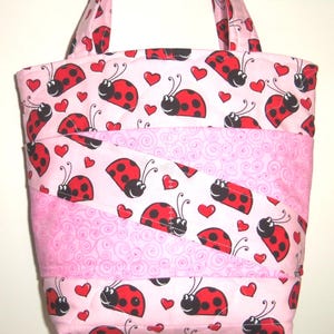 May include: A quilted tote bag featuring a repeating pattern of red and black ladybugs and small red hearts on a light pink background, interspersed with diagonal bands of a lighter pink fabric with a subtle swirling line pattern.