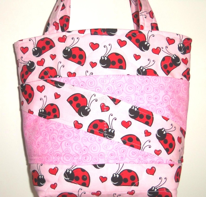 May include: A quilted tote bag featuring a repeating pattern of red and black ladybugs and small red hearts on a light pink background, interspersed with diagonal bands of a lighter pink fabric with a subtle swirling line pattern.