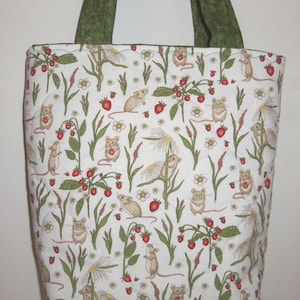 May include: A quilted tote bag featuring a white background with a repeating pattern of small brown mice, red strawberries, green leaves, and white daisy-like flowers. The bag has two dark green fabric handles attached to the top edge.