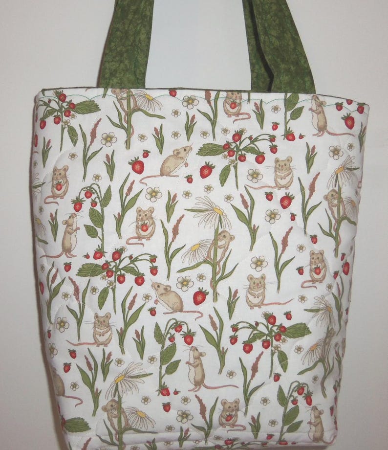 May include: A quilted tote bag featuring a white background with a repeating pattern of small brown mice, red strawberries, green leaves, and white daisy-like flowers. The bag has two dark green fabric handles attached to the top edge.