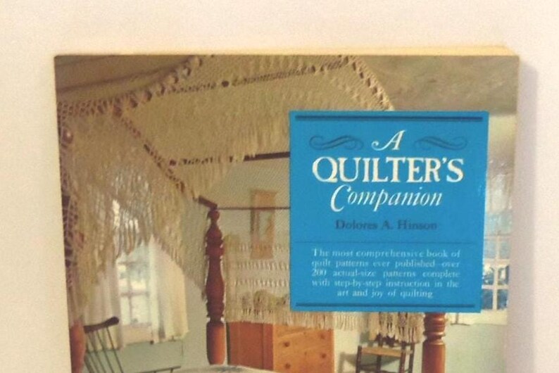 Puede incluir: Un libro titulado A Quilter's Companion de Doloris A. Hinson con una superposici&oacute;n rectangular azul y texto blanco. La portada muestra una cama con dosel con un dosel de ganchillo blanco y flecos en una habitaci&oacute;n con muebles de madera.