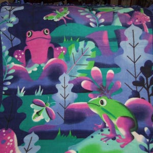 May include: A fleece blanket with a colourful frog and butterfly design in pink, green, purple, and blue. The blanket has a fringed edge in a darker blue. The design includes stylized plants and mushrooms.