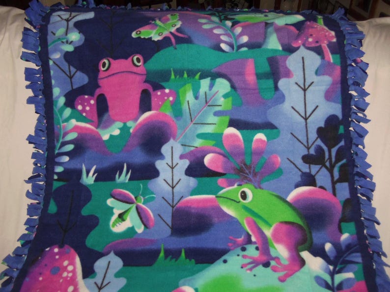 May include: A fleece blanket with a colourful frog and butterfly design in pink, green, purple, and blue. The blanket has a fringed edge in a darker blue. The design includes stylized plants and mushrooms.