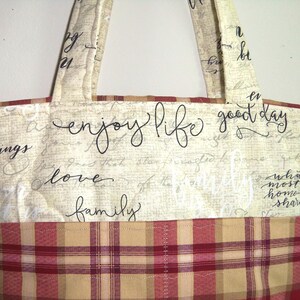 May include: A beige tote bag with a plaid fabric lining. The bag has black script text that reads "enjoy life good day love family".