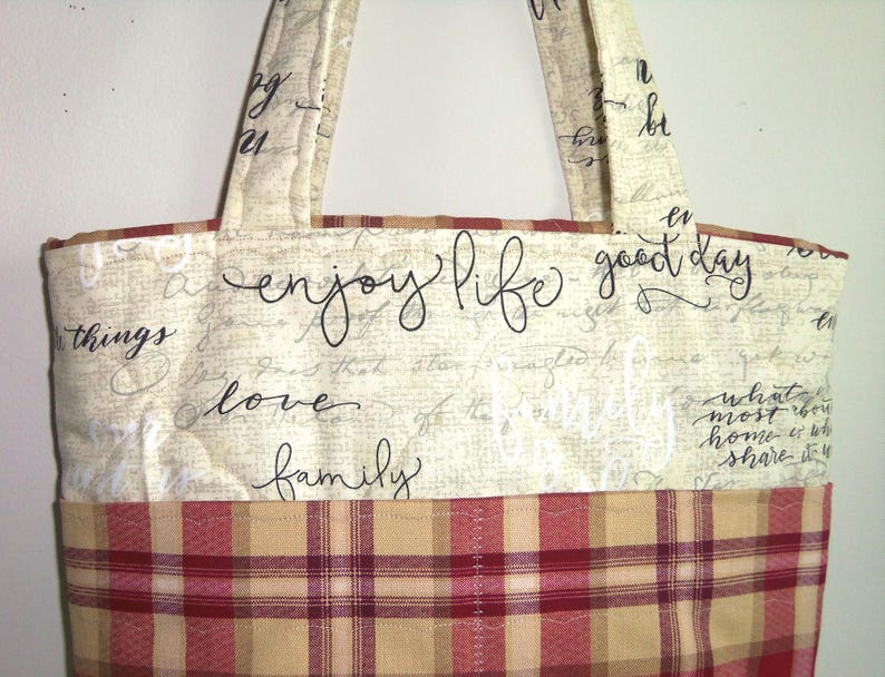 May include: A beige tote bag with a plaid fabric lining. The bag has black script text that reads "enjoy life good day love family".