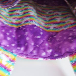 May include: A fabric tote bag featuring a quilted purple interior lining with a white heart pattern and an exterior section displaying a vibrant rainbow stripe design with white star accents, including two matching rainbow-patterned fabric handles.