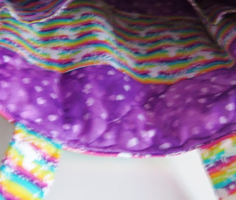 May include: A fabric tote bag featuring a quilted purple interior lining with a white heart pattern and an exterior section displaying a vibrant rainbow stripe design with white star accents, including two matching rainbow-patterned fabric handles.