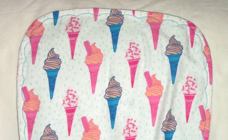 Op de afbeelding: Een witte stof met een geschulpte rand, met een herhalend patroon van ijsjes. De kegels zijn roze en blauw, met oranje en bruin ijs, sommige met hagelslag. Een blauwe gestikte rand.