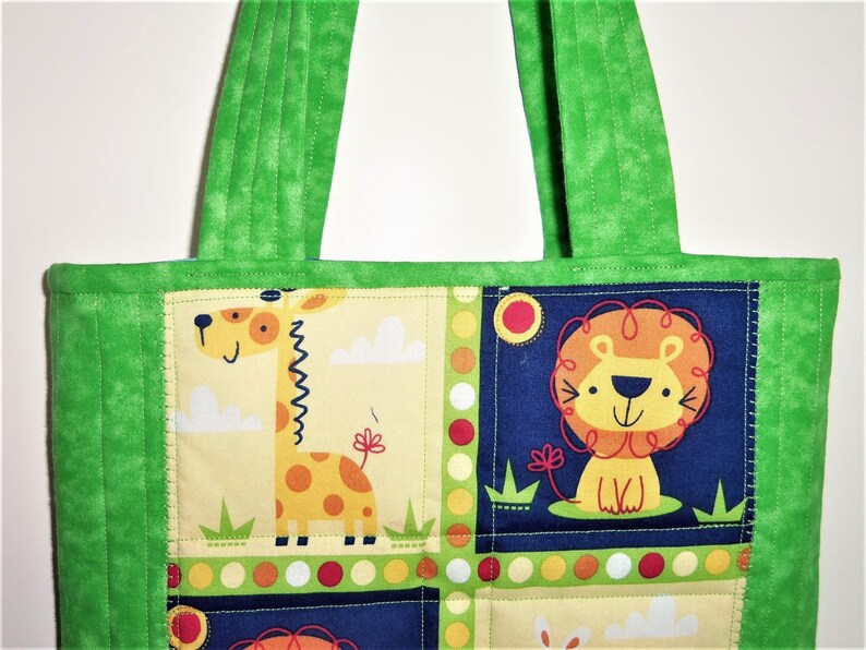 May include: A quilted tote bag featuring a vibrant green fabric border and handles. The central panel displays a patchwork design with cartoon illustrations of a yellow giraffe on a light background and a lion on a dark blue background.