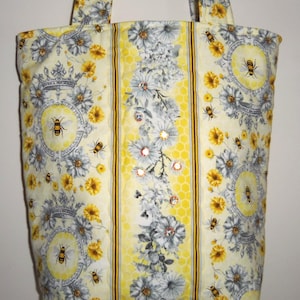 May include: A yellow tote bag with a bee and floral pattern. The bag features a repeating design of bees, flowers, and decorative elements. The center panel has a honeycomb pattern with floral accents and embellishments. The bag has two handles.