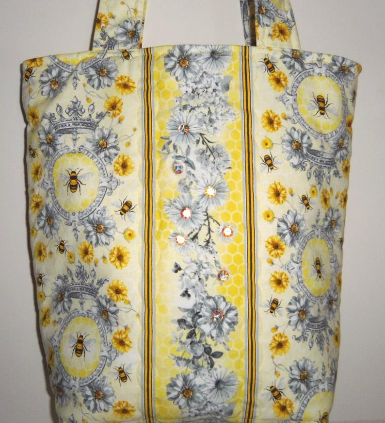 May include: A yellow tote bag with a bee and floral pattern. The bag features a repeating design of bees, flowers, and decorative elements. The center panel has a honeycomb pattern with floral accents and embellishments. The bag has two handles.