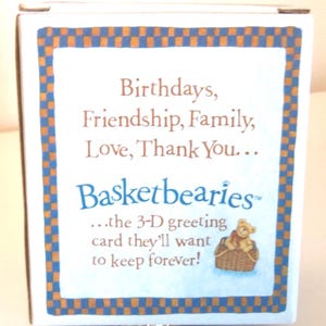 May include: A white box with a blue and orange checkered border. The text reads "Birthdays, Friendship, Family, Love, Thank You..." and "Basketbearies" with a small illustration of a teddy bear in a basket. The text continues, "...the 3-D greeting card they'll want to keep forever!"