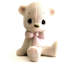 May include: A small, off-white ceramic bear figurine in a seated position. The bear features large, dark, teardrop-shaped eyes, a small green nose, and a light pink bow tie with subtle polka dots around its neck. The surface has a matte finish.