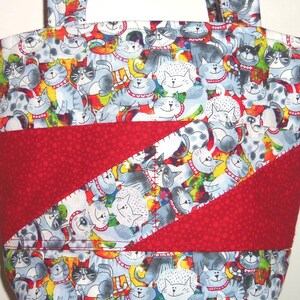 May include: A fabric tote bag featuring a repeating pattern of cartoon cats in shades of gray, white, and black with red collars. The bag includes a diagonal band of solid red fabric with a subtle tonal dot texture across the center front.