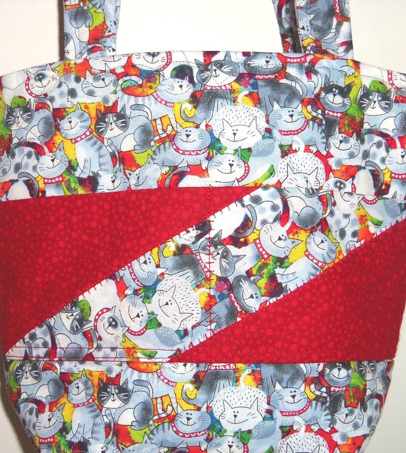 May include: A fabric tote bag featuring a repeating pattern of cartoon cats in shades of gray, white, and black with red collars. The bag includes a diagonal band of solid red fabric with a subtle tonal dot texture across the center front.