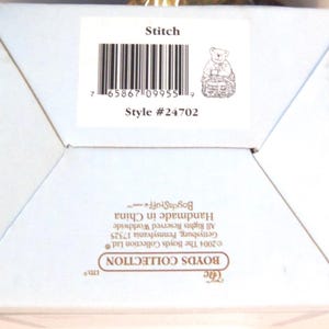 May include: A light blue box with the text "Stitch" and "Style #24702". The box features a small illustration of a bear holding a basket. The box also includes a barcode and the text "The Boyds Collection Ltd."