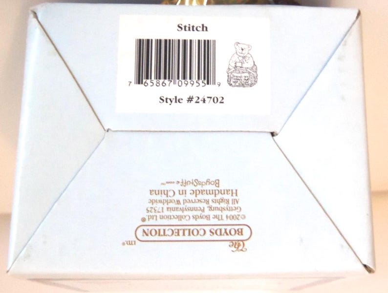 May include: A light blue box with the text "Stitch" and "Style #24702". The box features a small illustration of a bear holding a basket. The box also includes a barcode and the text "The Boyds Collection Ltd."
