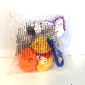 K&ouml;nnte beinhalten: Ein versiegelter, durchsichtiger Plastikbeutel mit einem kleinen Spielzeug, das eine leuchtend orangefarbene, abgerundete Basis, einen gelben Abschnitt und eine blaue Kunststoffschlaufe aufweist. Die Verpackung ist mit Sicherheitshinweisen bedruckt.